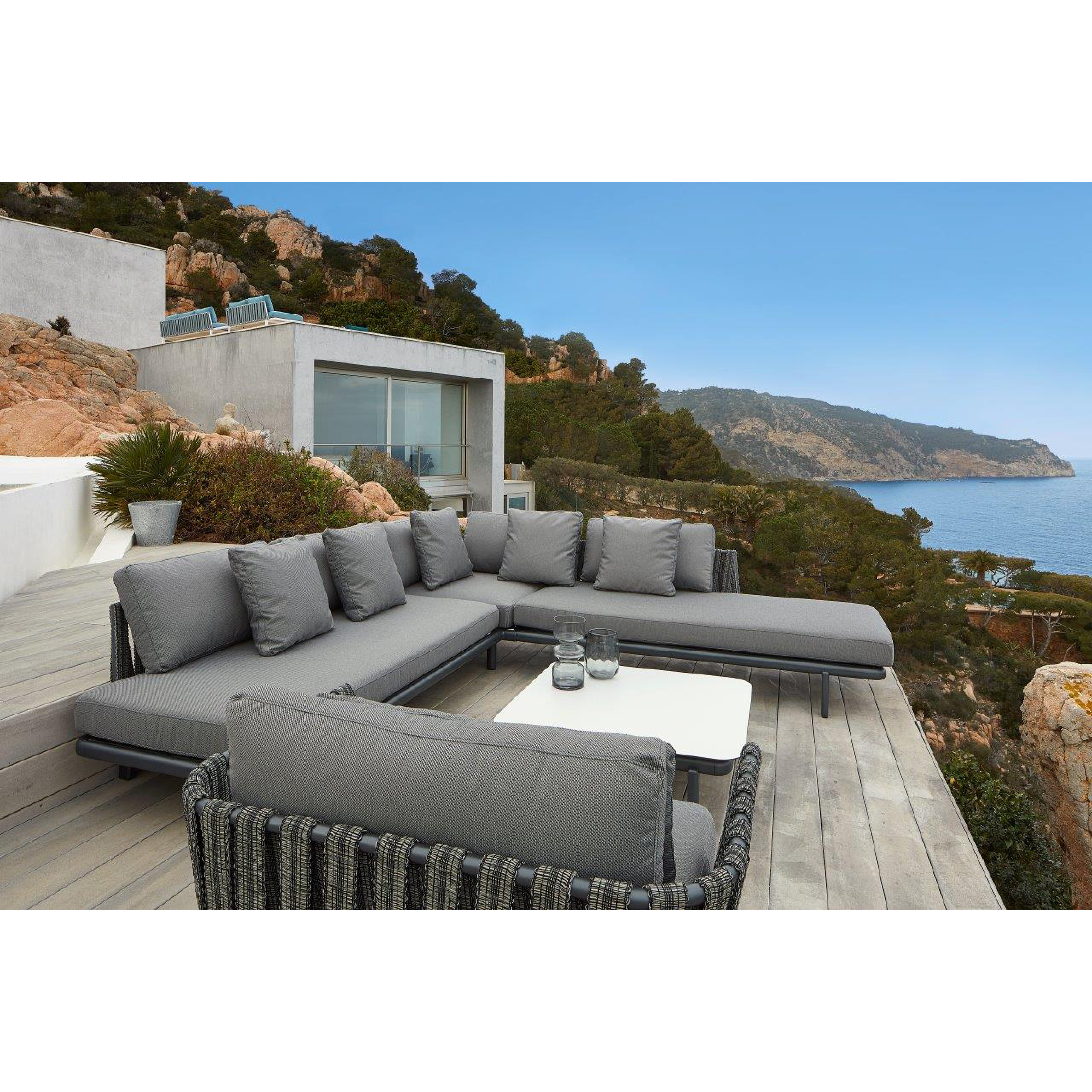 A modern outdoor patio features a Resol Anthea Sofa System and lounge chair on a wooden deck. A white table centers the scene, overlooking a scenic coastline with rocky hills and blue sea. Nearby, a contemporary house with glass windows complements the sleek aluminum structure of the furniture.