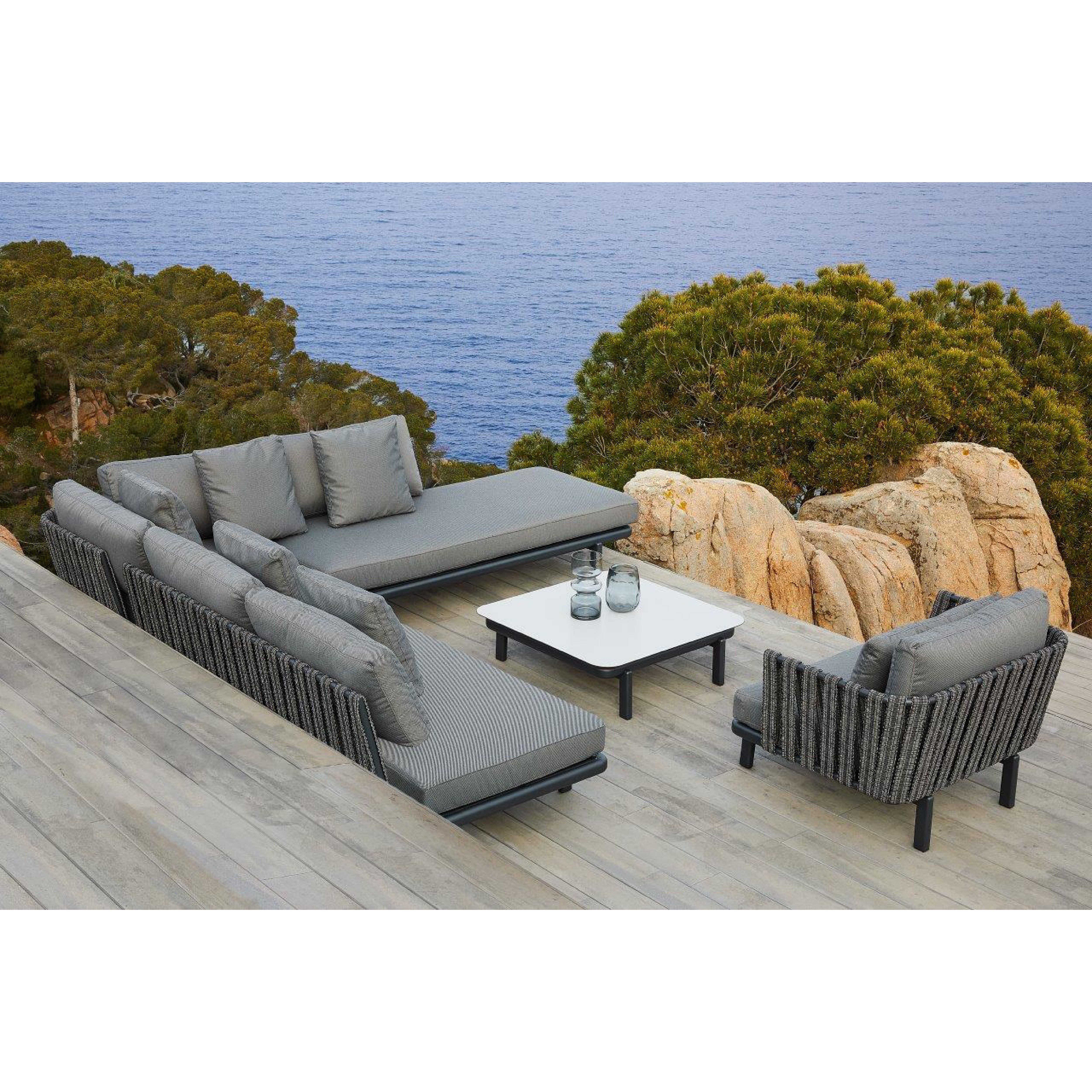 The patio boasts sleek outdoor furniture, featuring the Resol Anthea Sofa System with gray cushions and a low white table on a wooden deck. It offers an ocean view with rocky cliffs and is surrounded by lush greenery.