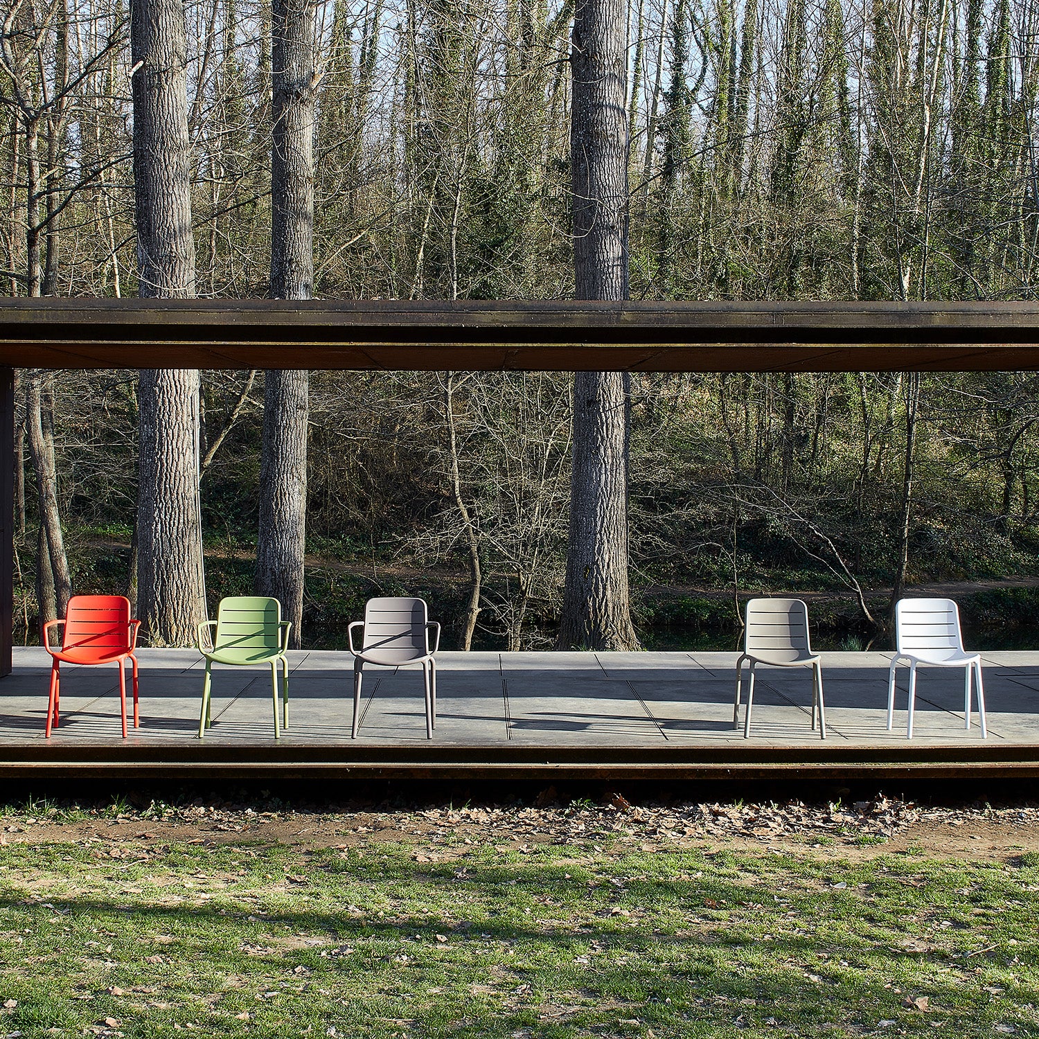 Five colorful Gina Armchairs by Resol—in red, green, yellow, beige, and gray—crafted sustainably, are arranged on a wooden platform against a lush backdrop of tall trees, providing an inviting and eco-friendly seating experience.