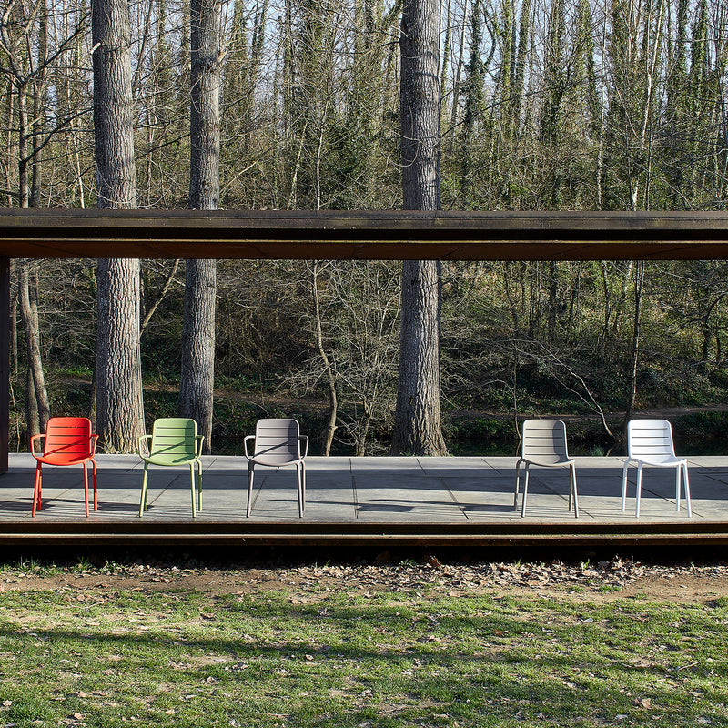 Five colorful Gina Armchairs by Resol—in red, green, yellow, beige, and gray—crafted sustainably, are arranged on a wooden platform against a lush backdrop of tall trees, providing an inviting and eco-friendly seating experience.