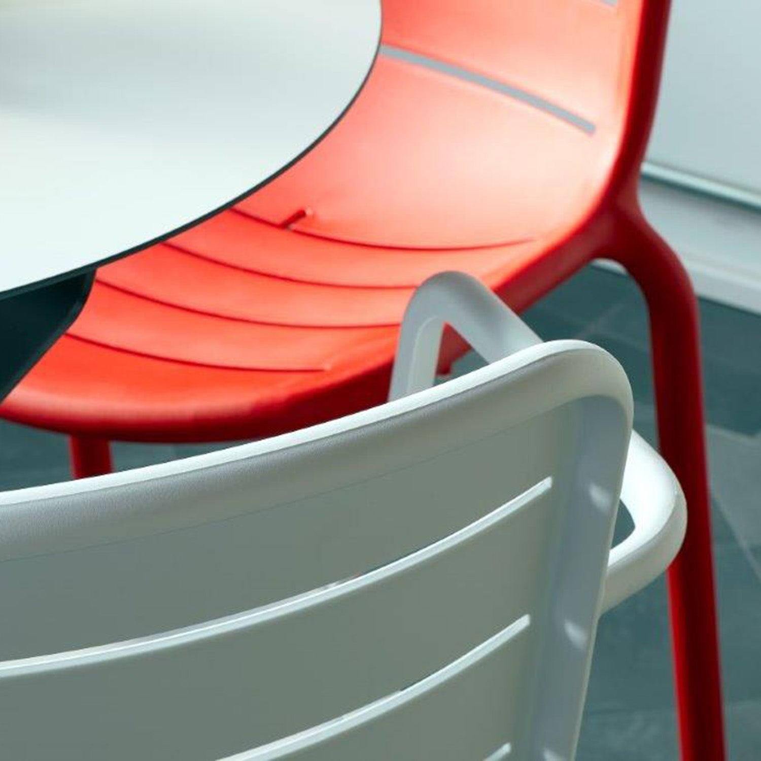 Close-up of two Gina Armchairs by Resol around a round table. This modern, innovative furniture features slatted backs; the partially visible chair is red and the more prominent one is white. Crafted sustainably, the scene rests on a smooth-finished dark floor.