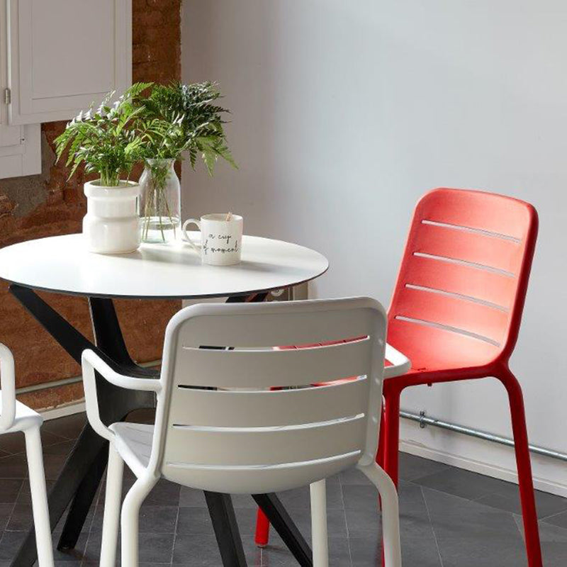 The setting features a round white table with two white chairs and one red Resol Gina Armchair on a dark tiled floor. Crafted sustainably, it includes a potted plant and mug on the table, set against brick and white walls.