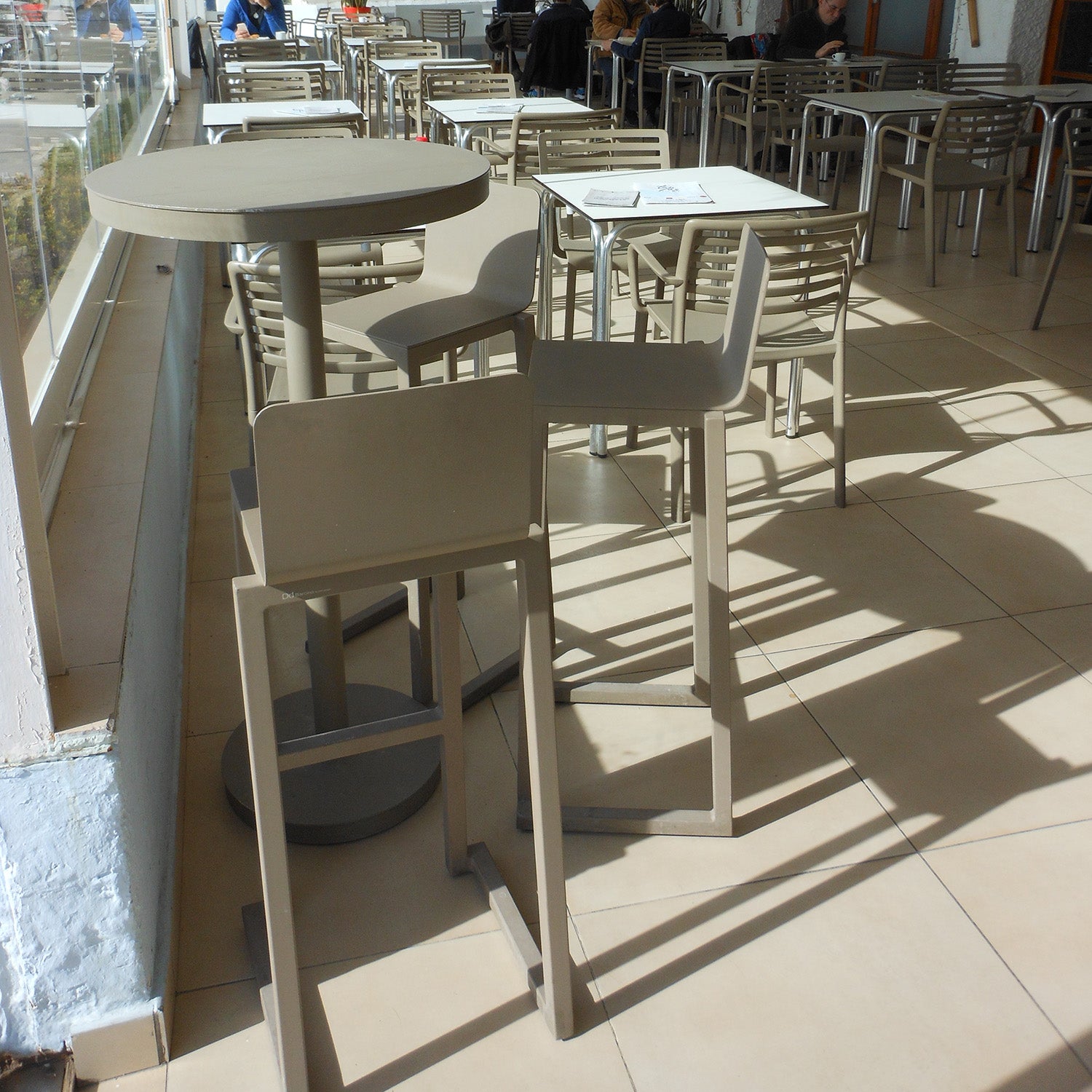 An empty café showcases beige tables and chairs made from powder-coated aluminum, sunlight casting shadows on the tiled floor. A few people sit near the glass windows, enjoying the sustainable Barcino Barstool by Resol, designed by Joan Gaspar.
