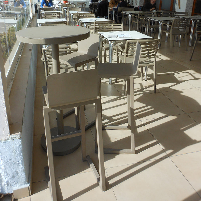 An empty café showcases beige tables and chairs made from powder-coated aluminum, sunlight casting shadows on the tiled floor. A few people sit near the glass windows, enjoying the sustainable Barcino Barstool by Resol, designed by Joan Gaspar.