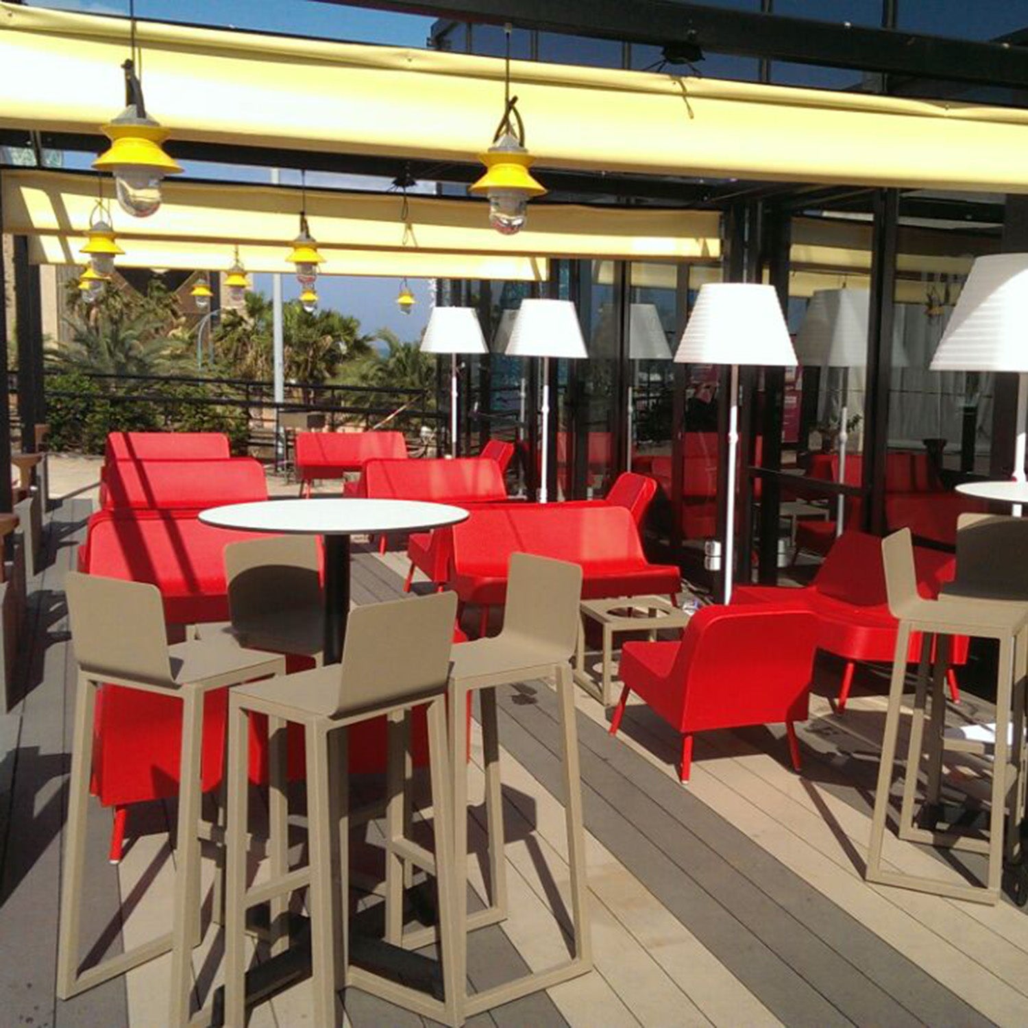 A modern patio showcases sustainable red and beige furniture, featuring Resols Barcino Barstool made of powder-coated aluminum. Red chairs and sofas complement tables, under a yellow canopy that shades the space adorned with suspended yellow lamps and greenery for a relaxed ambiance.