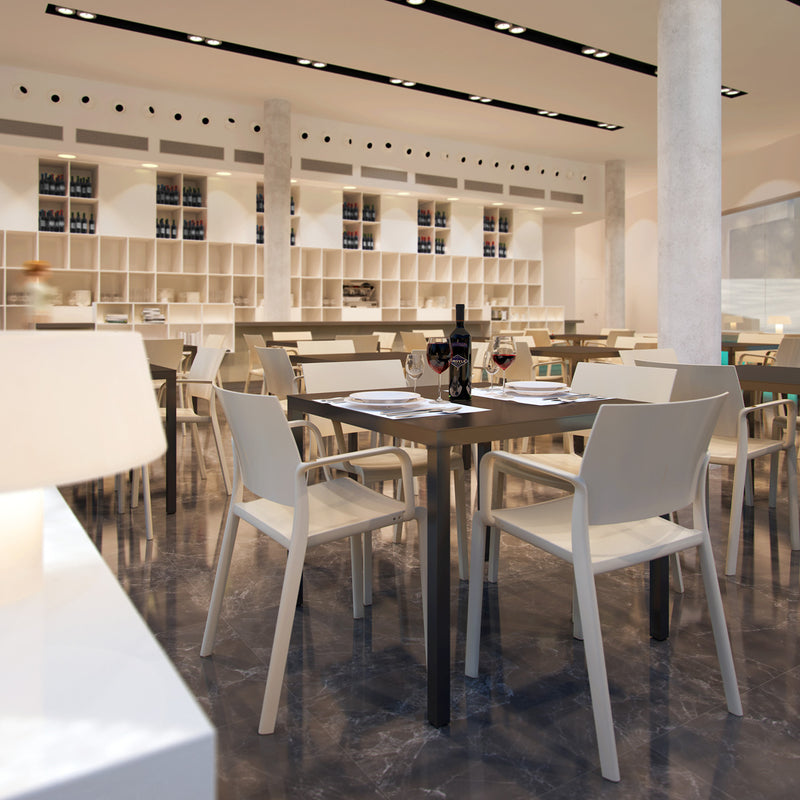 A modern restaurant interior includes Resols Barcino Barstools and white chairs crafted from sustainable furniture on a polished floor. Tables feature wine glasses and a bottle of wine, while shelves with various bottles adorn the back wall, all enhanced by ceiling lights.