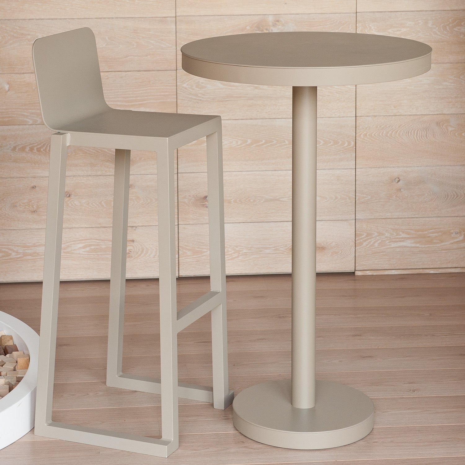 The Resol Barcino Barstool, with minimalistic elegance in beige and sustainable design, stands tall beside a matching round bar table. Set against light wood paneling and powder-coated aluminum accents, it enhances the warm ambience in this modern setting.