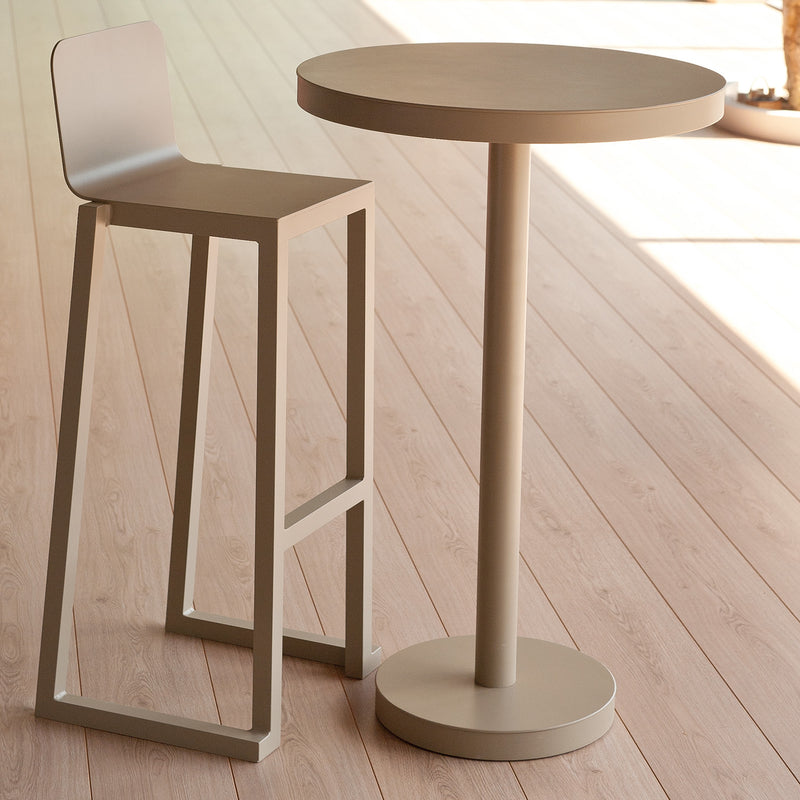In a minimalist indoor space, the Resol Barcino Barstool and a round table of powder-coated aluminum bring elegance. The tables pedestal base complements its smooth design, while natural light highlights the sustainable furniture and wooden floor, enhancing the serene ambiance.