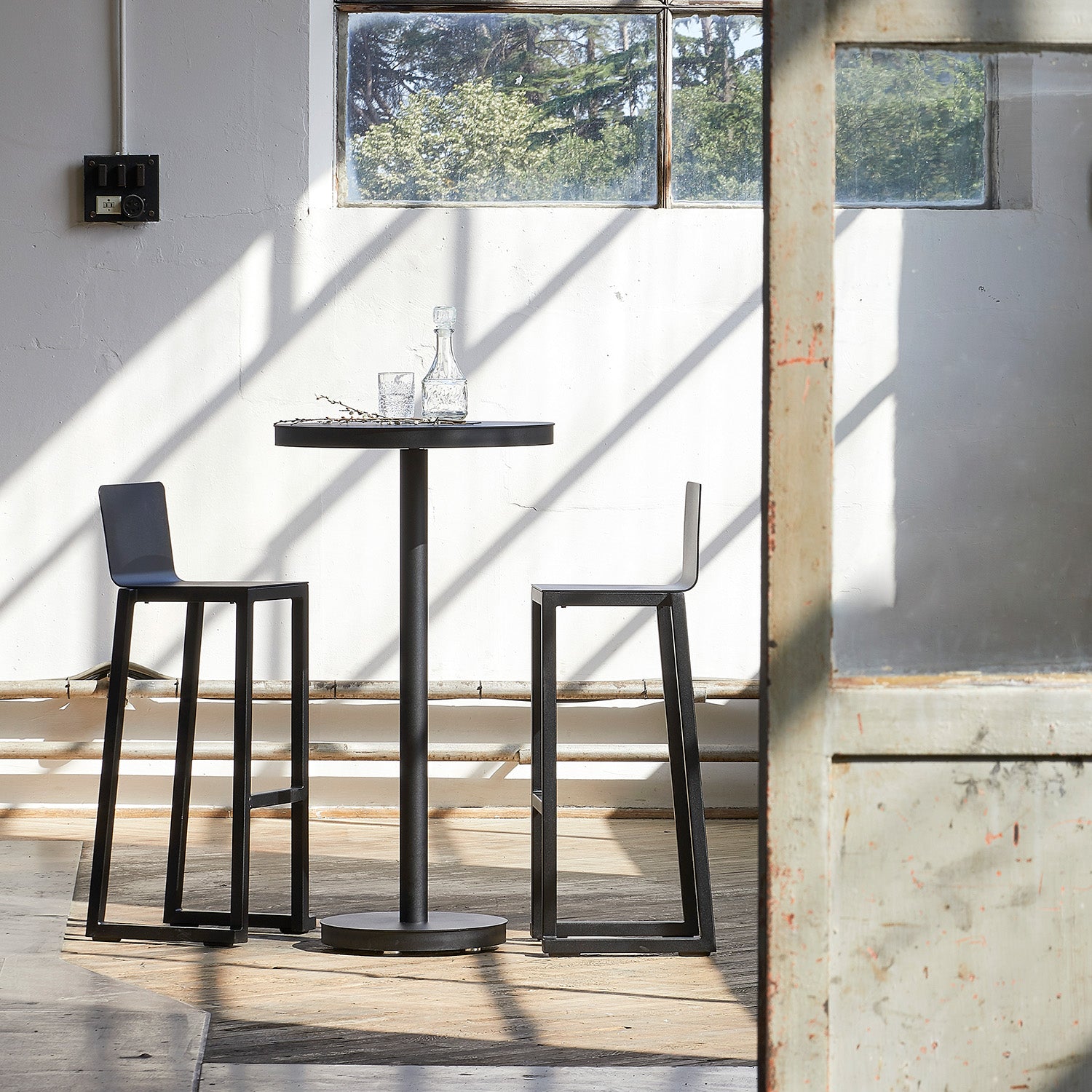 A small round table with two Resol Barcino Barstools, crafted from powder-coated aluminum, sits in a sunlit room. A glass decanter and two glasses rest on the table as sunlight casts window frame shadows on white walls. A large window and partially open wooden door complete the scene.