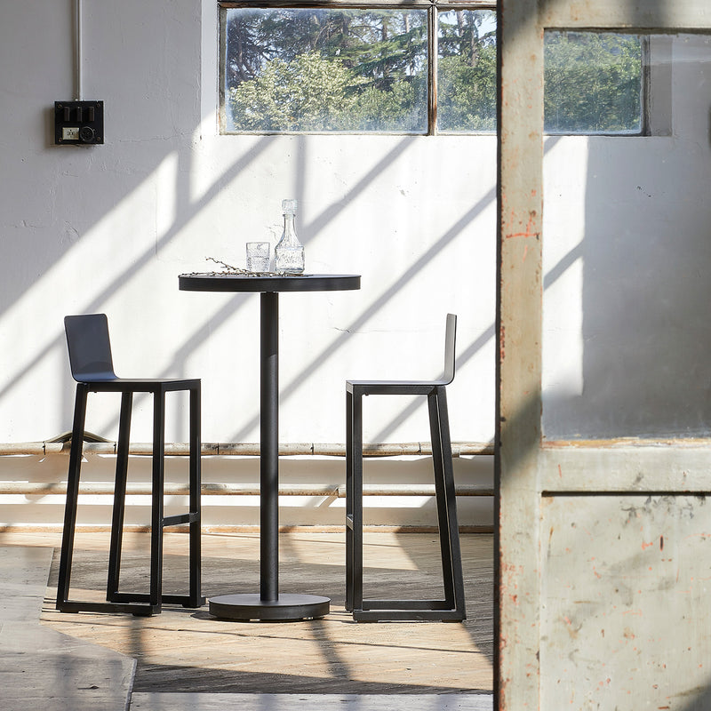 A small round table with two Resol Barcino Barstools, crafted from powder-coated aluminum, sits in a sunlit room. A glass decanter and two glasses rest on the table as sunlight casts window frame shadows on white walls. A large window and partially open wooden door complete the scene.