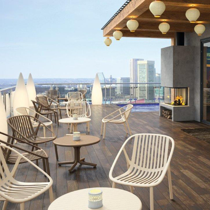 Rooftop patio with Resols modern Bini Armchairs and tables on wooden decking, overlooking a city skyline. Lanterns hang from the awning, with a decorative fireplace on the side. The view includes tall buildings and a river in the background. UV protection ensures durability for outdoor settings.