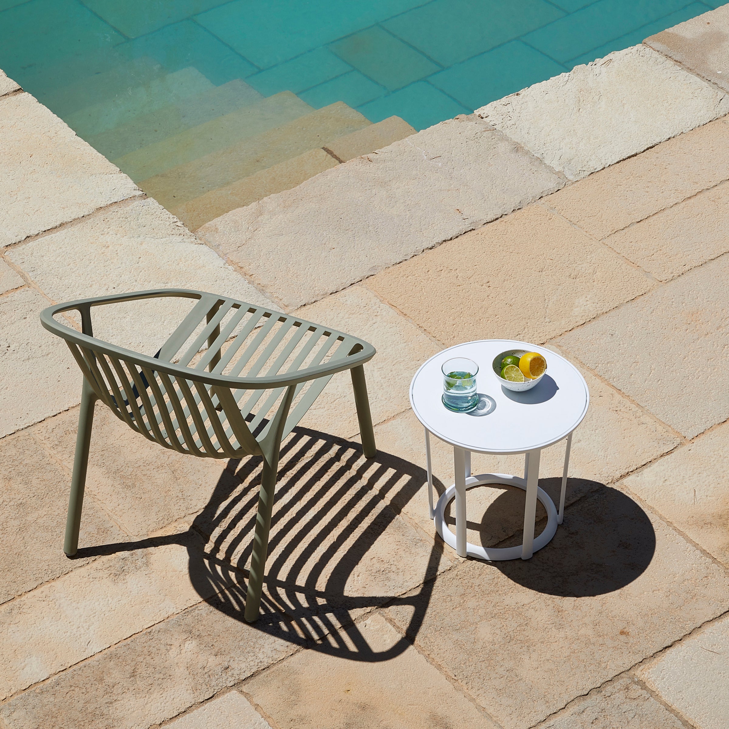 The Bini Armchair by Resol, sustainably crafted in green, is placed beside a round white table holding a glass of water and a bowl of lemons on a stone patio next to the turquoise-tiled pool. Shadows from the UV-protected chair dance gracefully on the patio.