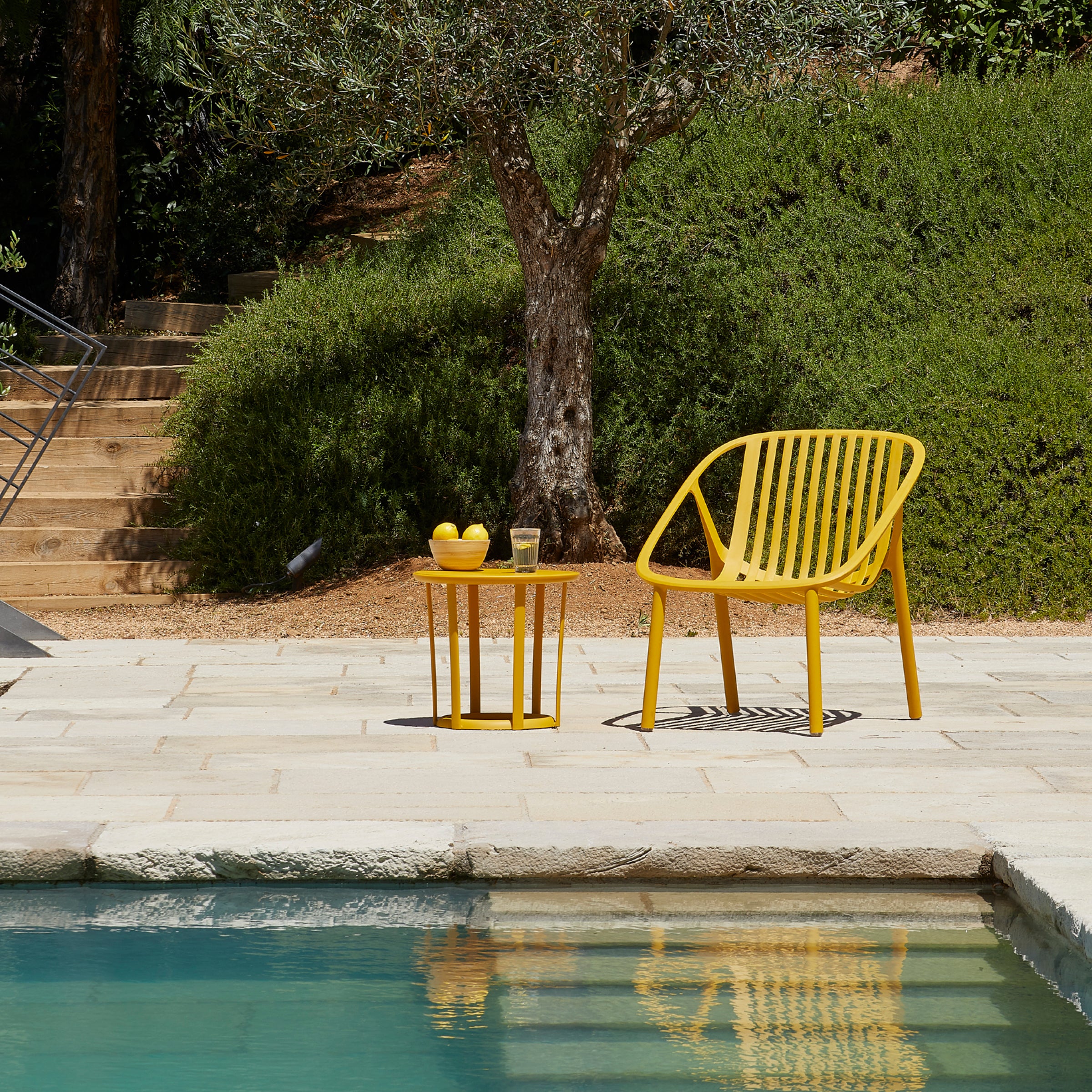 A Bini Armchair in yellow by Resol and a side table set with lemons and a glass grace the stone patio by the swimming pool. A large bush and tree enhance the background, with steps leading up a grassy hill. Crafted through sustainable production.