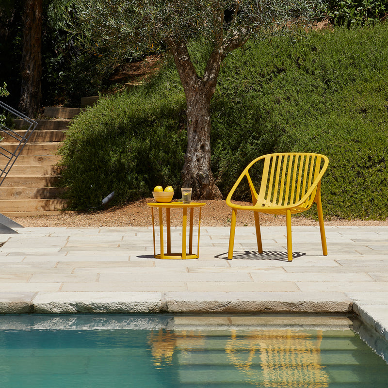 A Bini Armchair in yellow by Resol and a side table set with lemons and a glass grace the stone patio by the swimming pool. A large bush and tree enhance the background, with steps leading up a grassy hill. Crafted through sustainable production.