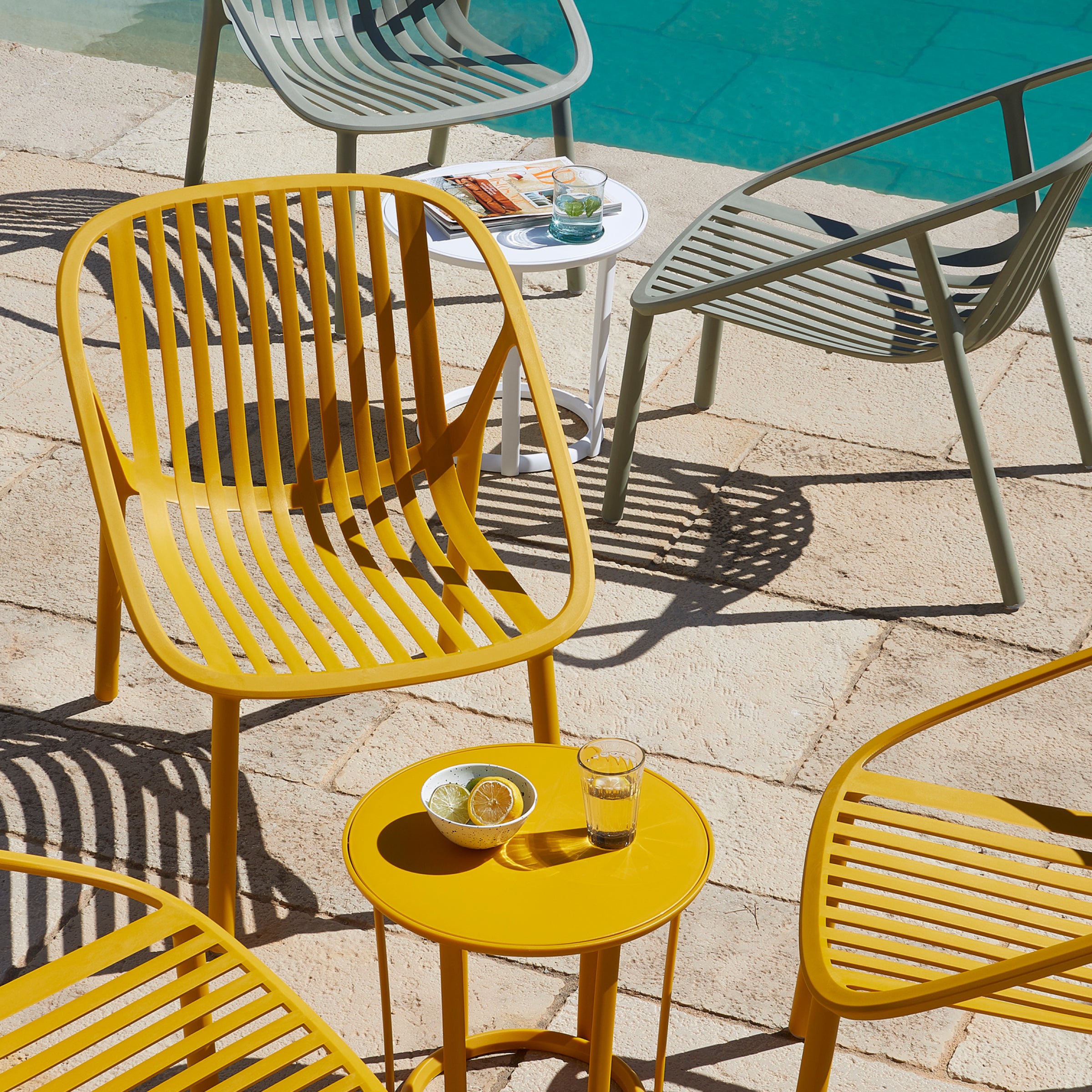 UV-protected yellow and green Bini Armchairs by Resol, paired with small round tables, adorn a stone patio with a pool in the background. One table features lemons and water, another holds a magazine and sunglasses. Shadows form patterns on the ground.