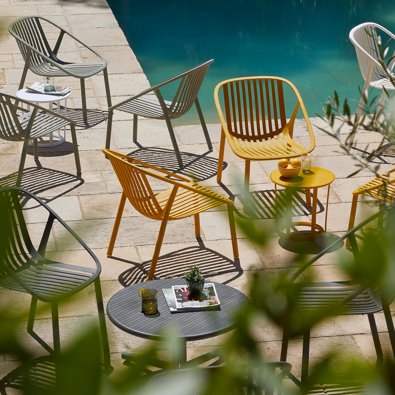 The outdoor patio scene showcases colorful, UV-protected Bini Armchairs by Resol and small tables near a pool. Yellow, white, and gray seating enhance the relaxed vibe. A magazine and drinks are on the tables, while green foliage elegantly frames the scene, highlighting these smart furniture solutions.