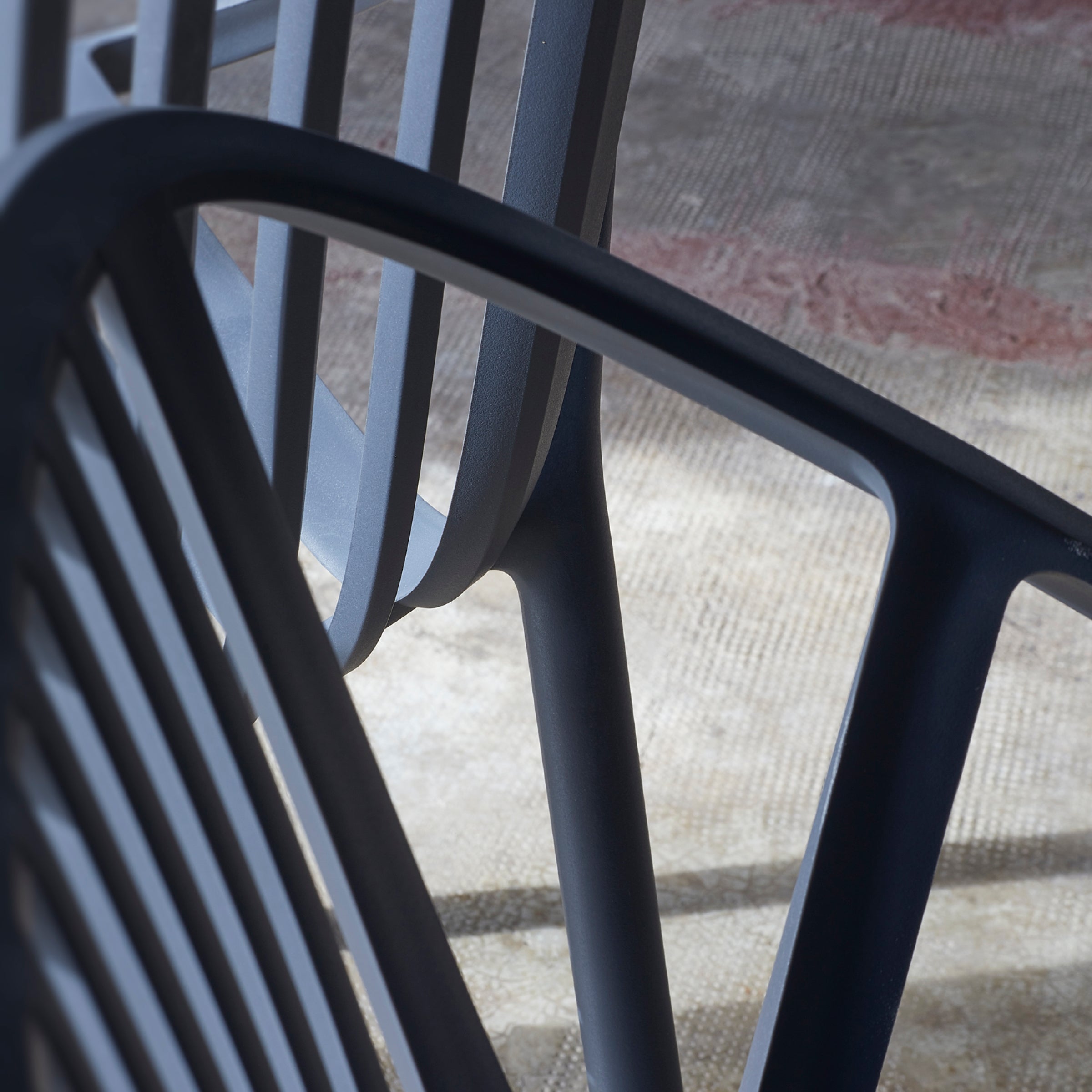 Close-up of overlapping Bini Armchair backs by Resol on a textured brown and beige floor, highlighting sustainable design. The black metal chairs cast subtle shadows, creating abstract patterns with lines, shapes, and angles.