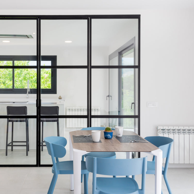 The modern dining area includes sustainable furniture like a wooden table and four Resol Casino Chairs in blue. A glass sliding door opens to the kitchen with a large window and black bar stools. The minimalistic space is bright, emphasized by white walls and a black-framed door.