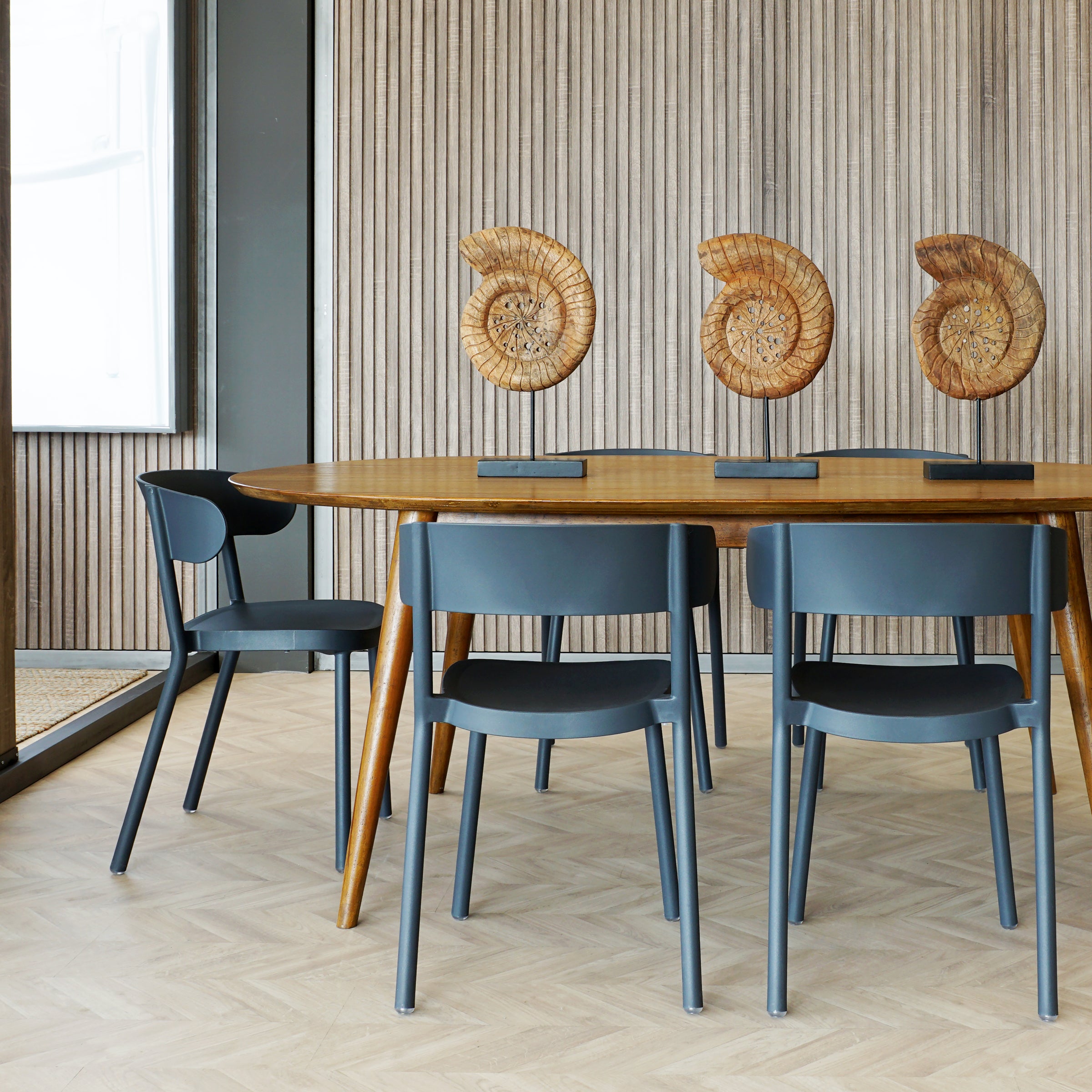 Modern dining area with a sustainable wooden table and four dark gray Casino Chairs by Resol. Three spiral fossil sculptures adorn the table, set against a textured wooden wall. Light wooden parquet flooring underscores the minimalist design, blending elegance with eco-friendly living.