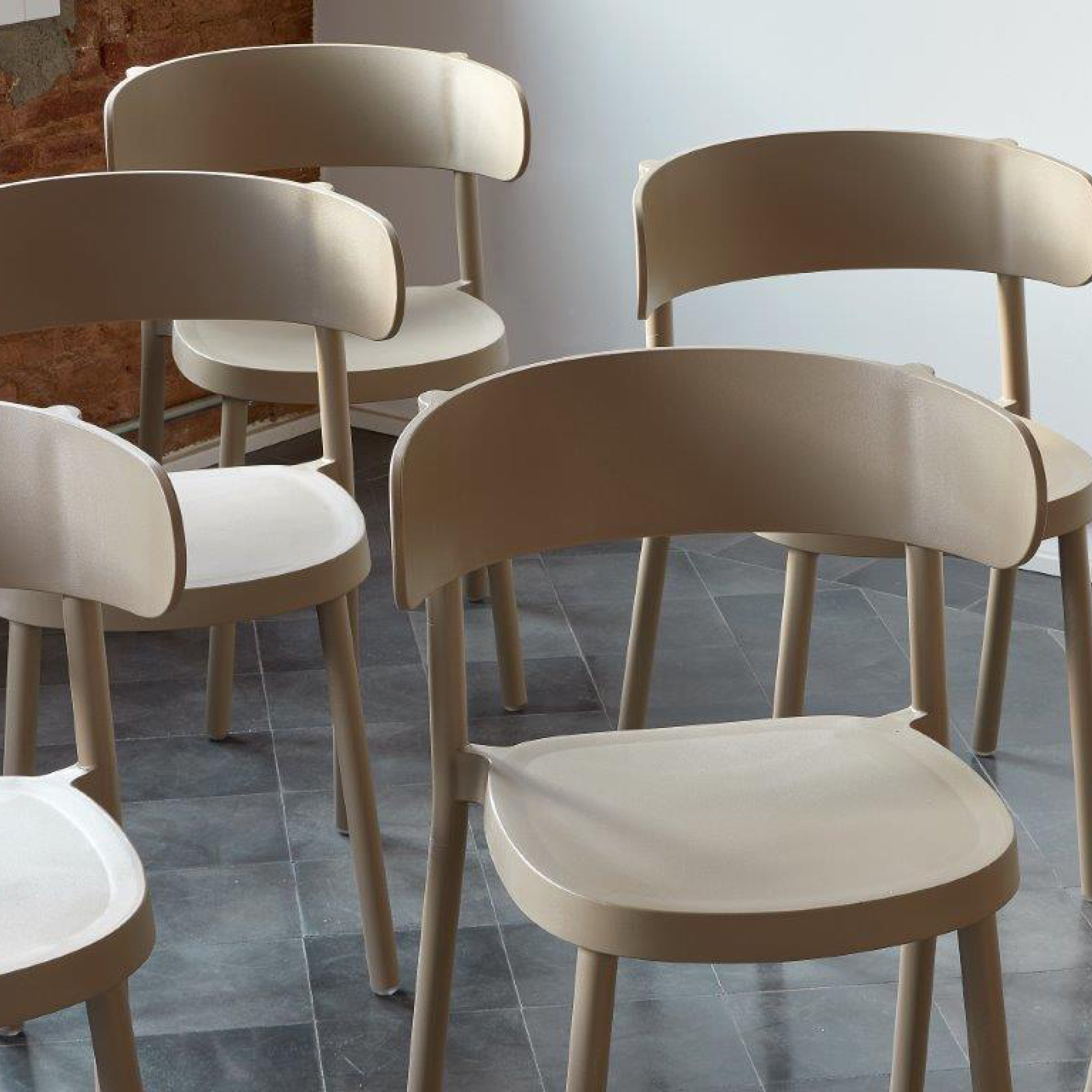 A group of light beige Resol Casino chairs with curved backs is arranged on a dark tiled floor. The background features a white wall and exposed brick, with natural light highlighting the sustainable choice and stylish setting.