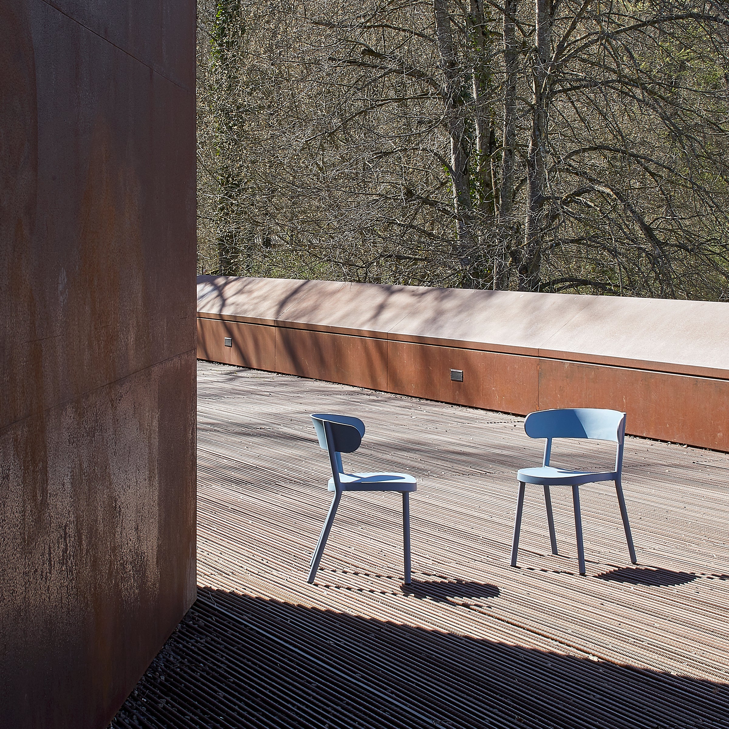 Two Resol Casino Chair UV-protected blue chairs sit on a wooden deck, with rust-colored walls to the left and trees in the background. The setting is calm and minimalist, softly illuminated by natural light.