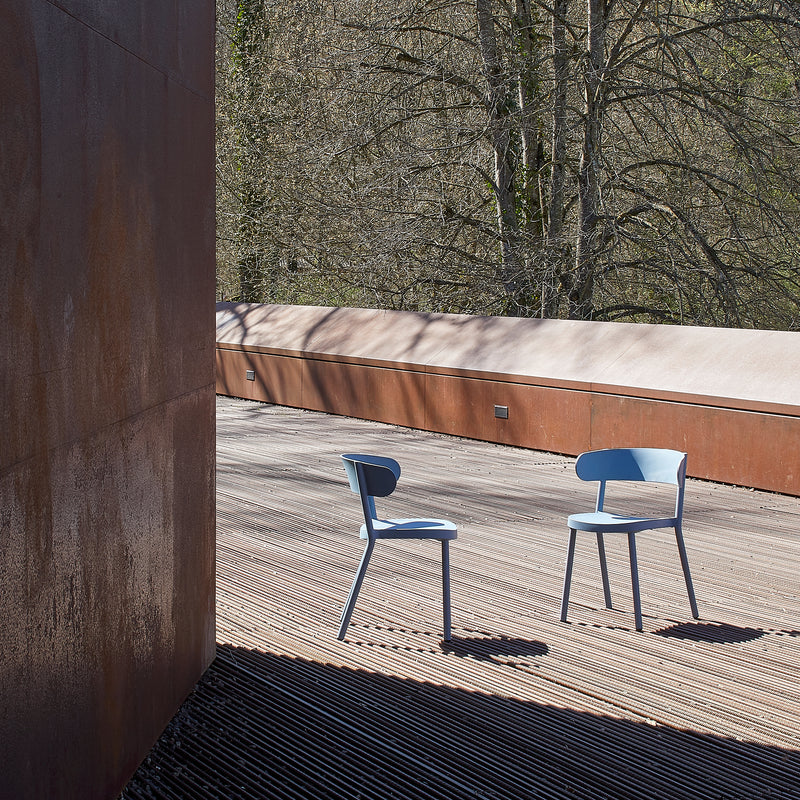 Two Resol Casino Chair UV-protected blue chairs sit on a wooden deck, with rust-colored walls to the left and trees in the background. The setting is calm and minimalist, softly illuminated by natural light.