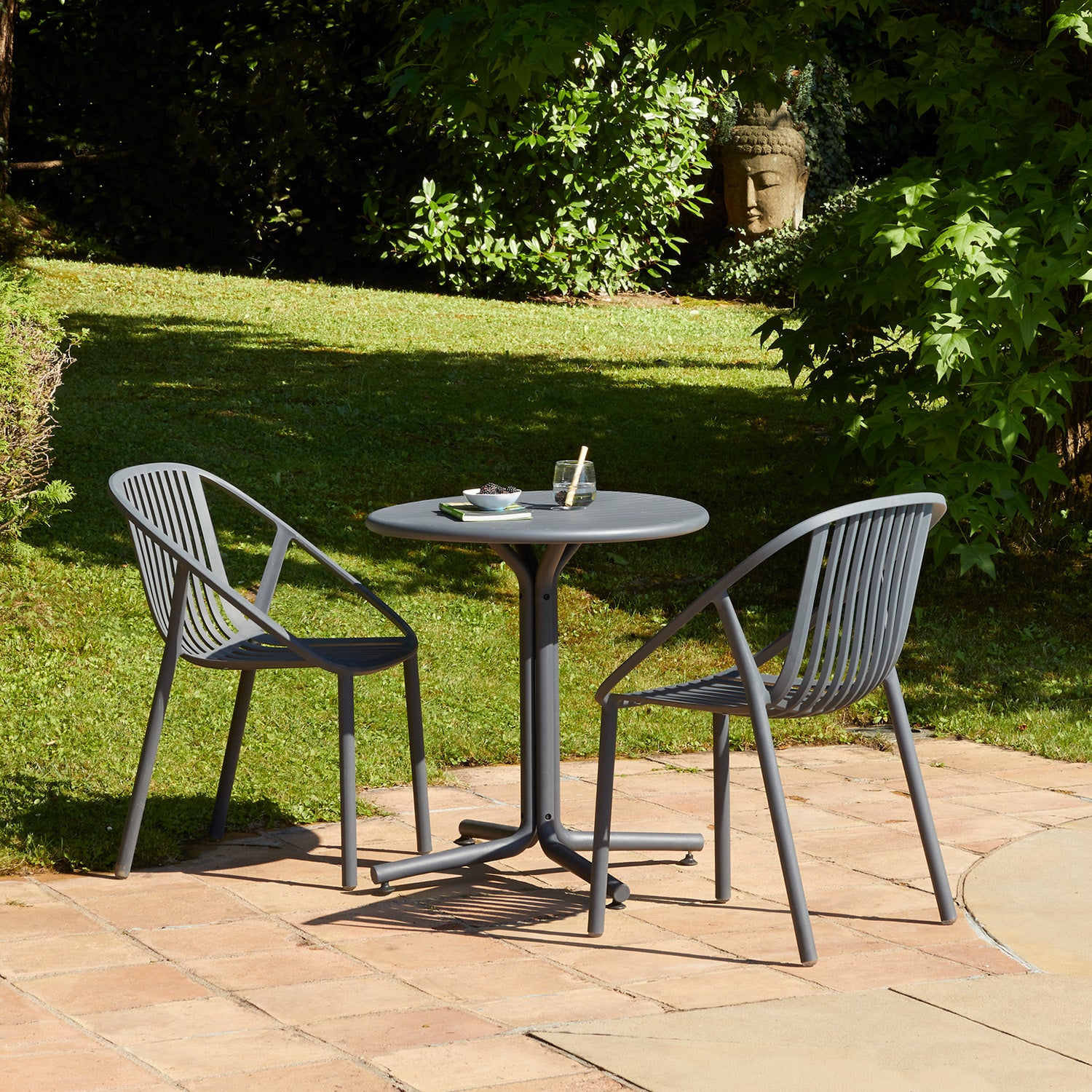 A serene garden setting features a small round table and two Resol Bini Chairs in UV-protected gray on a tiled patio. The table holds a green ashtray and glass, while lush greenery and a stone Buddha head bask in sunlight in the background.
