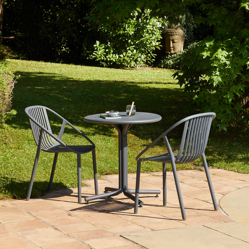 A serene garden setting features a small round table and two Resol Bini Chairs in UV-protected gray on a tiled patio. The table holds a green ashtray and glass, while lush greenery and a stone Buddha head bask in sunlight in the background.