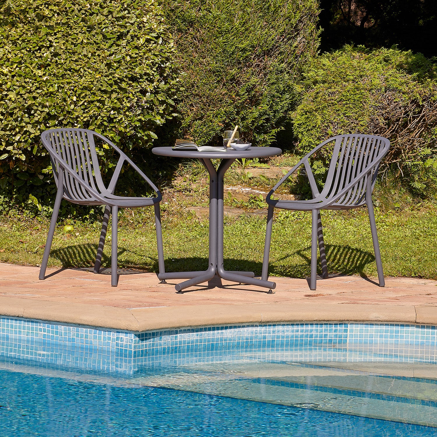 Two Resol Bini gray chairs and a round table, topped with a book and glass, are set on a stone patio beside a blue-tiled pool. UV-protected materials ensure durability, while lush green bushes and grass create a serene outdoor backdrop.