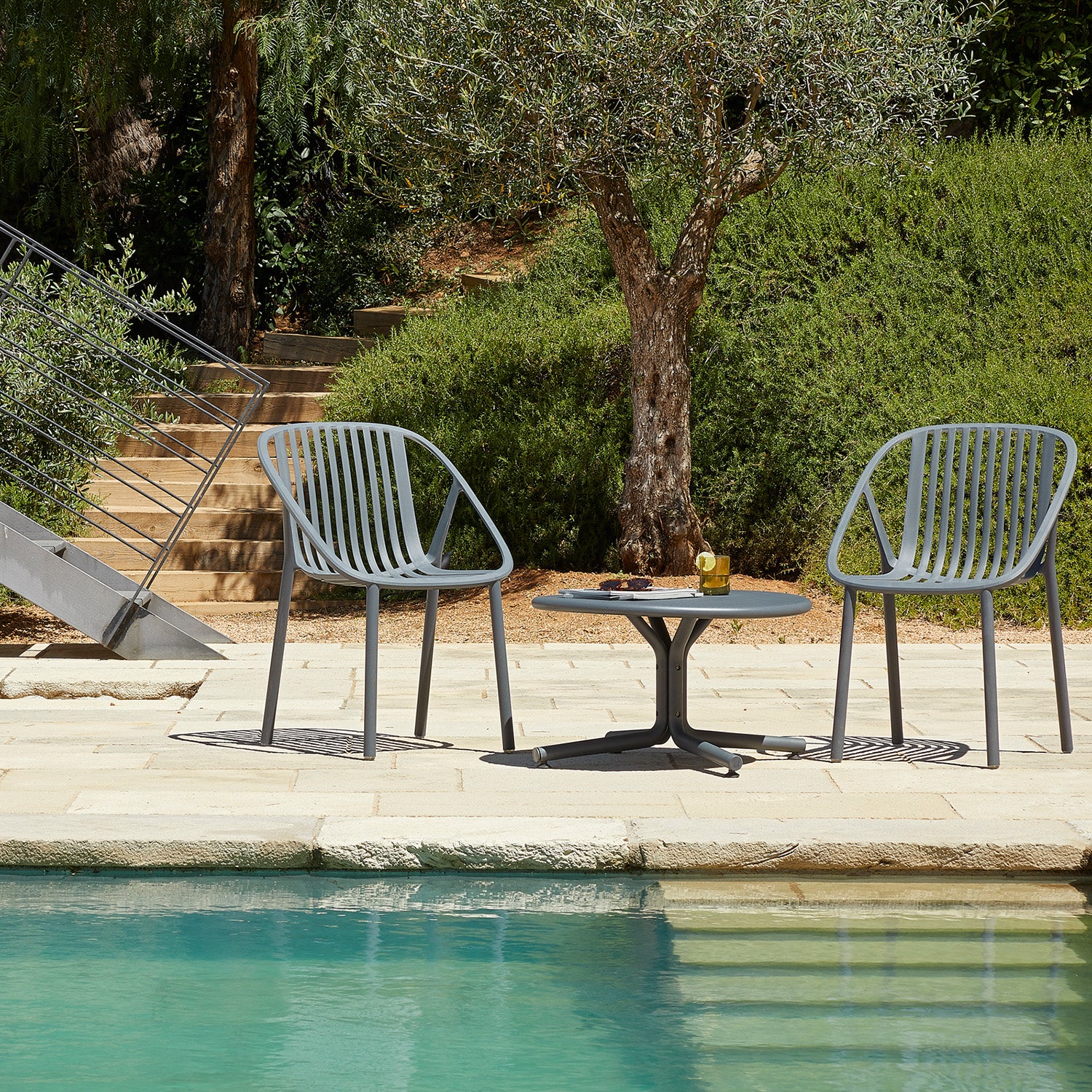 A pair of Resol Bini Chairs, crafted sustainably, sit by a pool with a small table holding a drink with lime. Sunlit skies highlight the lush greenery and an olive tree in the background.