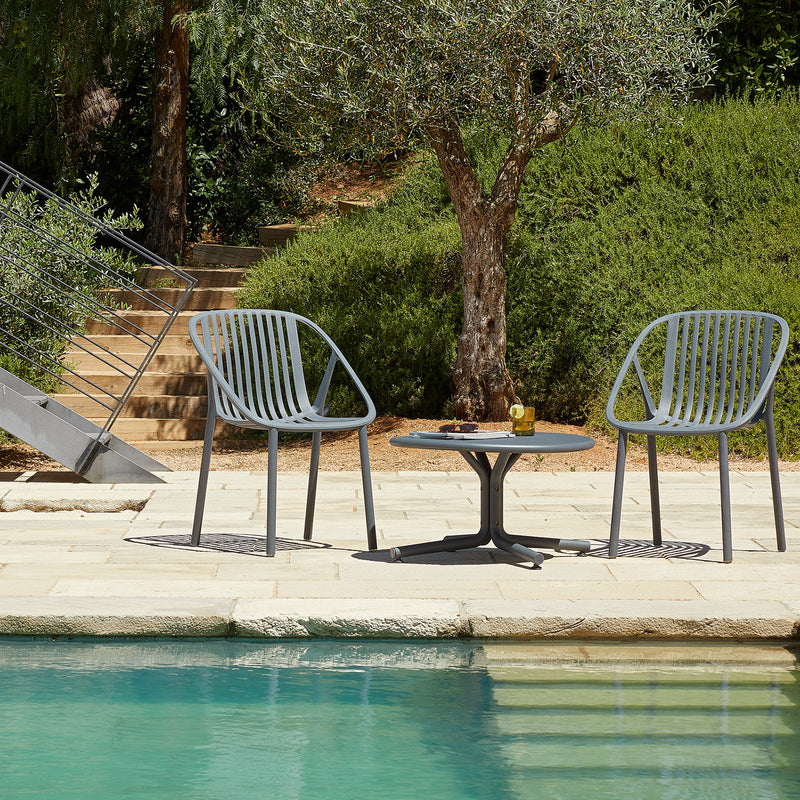 A pair of Resol Bini Chairs, crafted sustainably, sit by a pool with a small table holding a drink with lime. Sunlit skies highlight the lush greenery and an olive tree in the background.