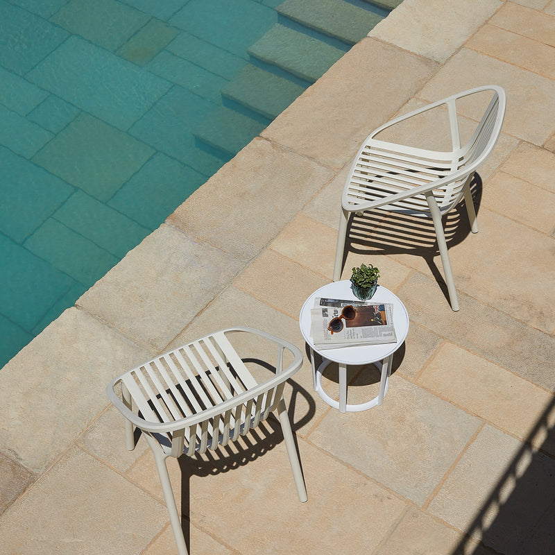 Two modern white Bini Chairs by Resol are placed beside a blue pool on a stone patio. A small round table with a magazine and potted plant sits between them, casting shadows on the ground, creating an inviting spot for peaceful outdoor relaxation. Crafted sustainably.