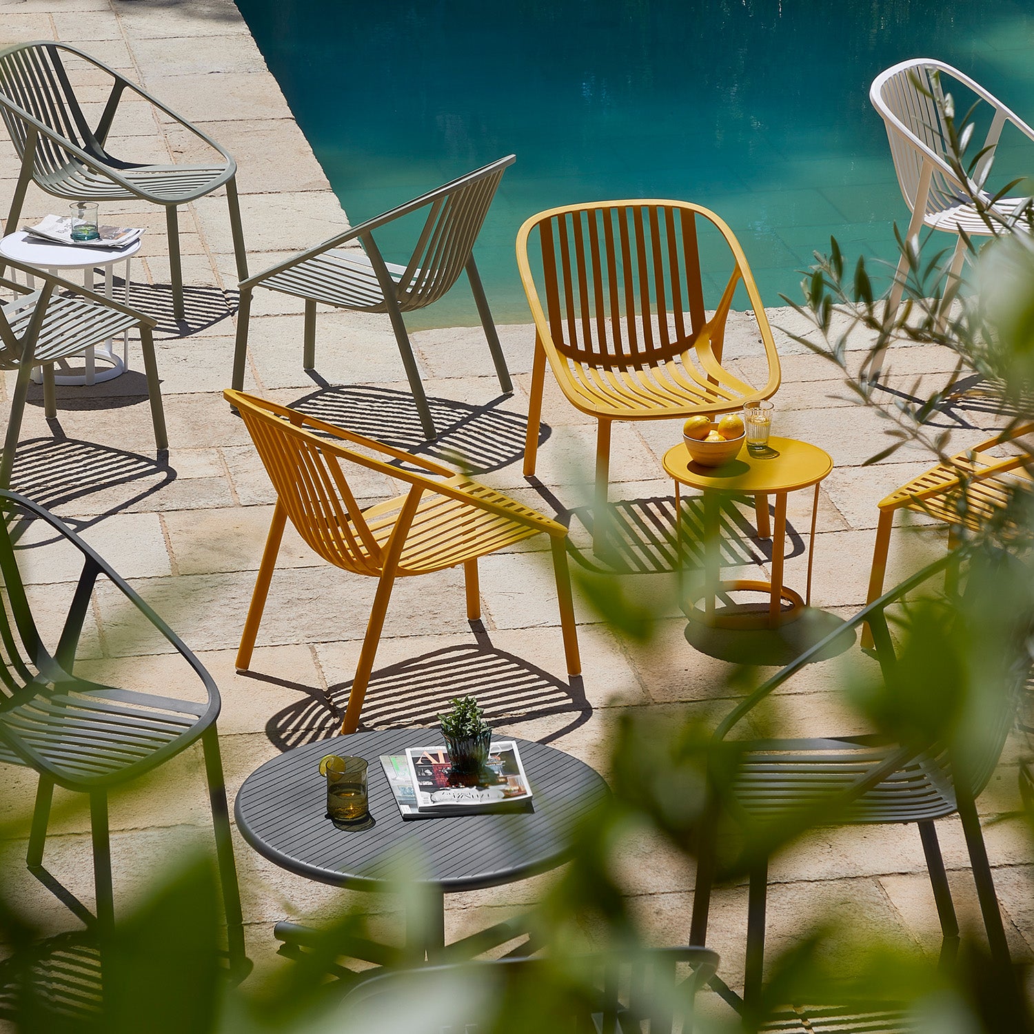 A scenic patio showcases innovative furniture with Resols Bini Chairs in modern yellow, gray, and white—all UV protected—overlooking a tranquil pool. Tables hold drinks and a magazine, while greenery frames the scene, enhancing the serene atmosphere.