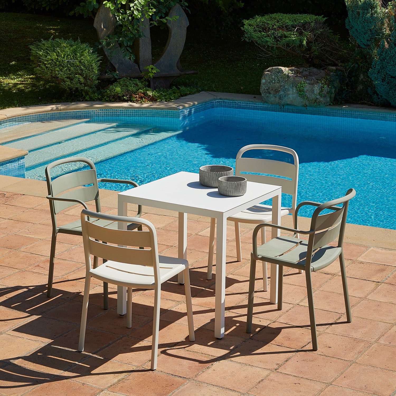 A white patio table with four Como Armchairs from Resol, crafted sustainably, sits on a terracotta-tiled patio by a turquoise pool. Two gray bowls are on the table, while greenery and an elegantly carved stone sculpture enhance the serene backdrop.