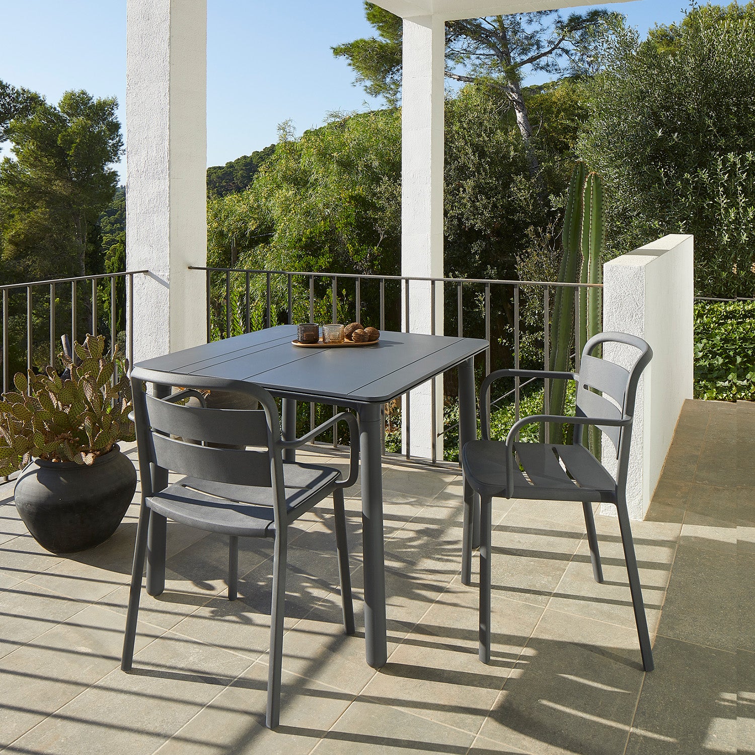 The sunny patio features innovative furniture with Resols Como Armchair and a modern outdoor table. A UV-protected tabletop holds a wooden tray with cups, while a large potted cactus is on the left, surrounded by lush greenery beyond the railings.