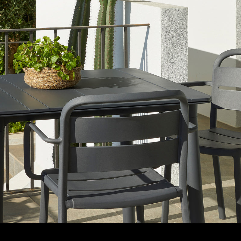 An outdoor patio setup includes a Como Armchair from Resol, paired with a dark grey table. A woven basket containing a green plant sits on the table, while shadows play on a white wall in the sunlight behind a large cactus.