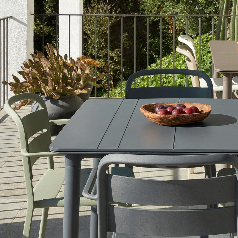 A patio highlights a gray table with a wooden bowl of apples, surrounded by Resols innovative Como Armchairs in UV-protected gray and green. Nearby, theres a railing, a large pot with a cactus, plus greenery and trees in the background.