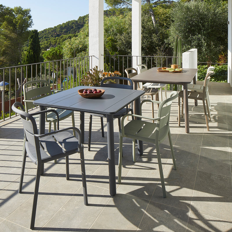 Two outdoor dining tables with Resols Como Armchairs are placed on a spacious balcony, one featuring a bowl of fruit. Crafted sustainably, this scene is enveloped by greenery and trees, creating a tranquil atmosphere under the sunny sky.
