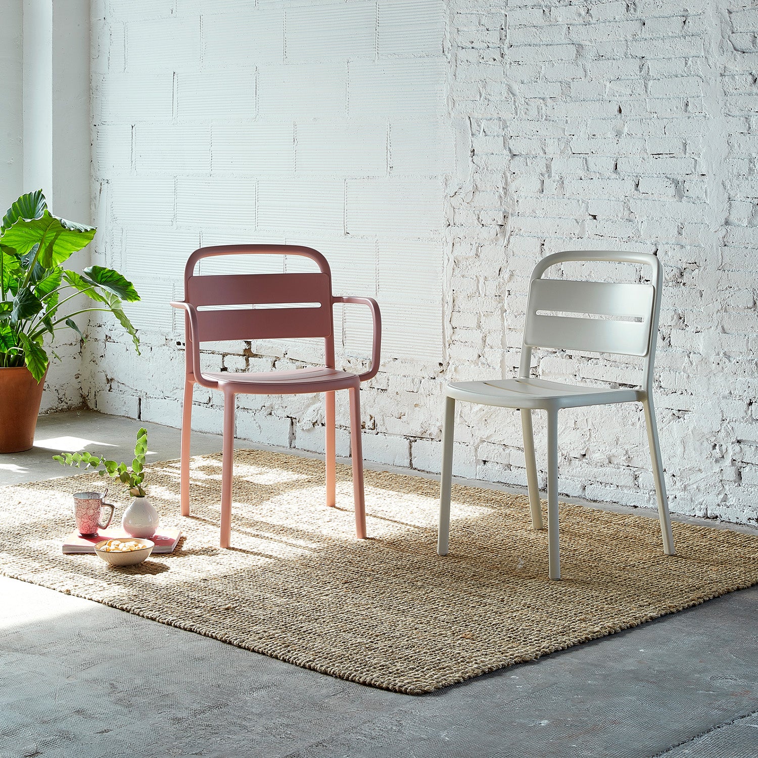 Two Resol Como Armchairs, in pink and beige, crafted sustainably, sit on a woven rug against white brick walls. Nearby are a potted plant and a small table with snacks and a glass jug. Sunlight from a large window brightens the room.