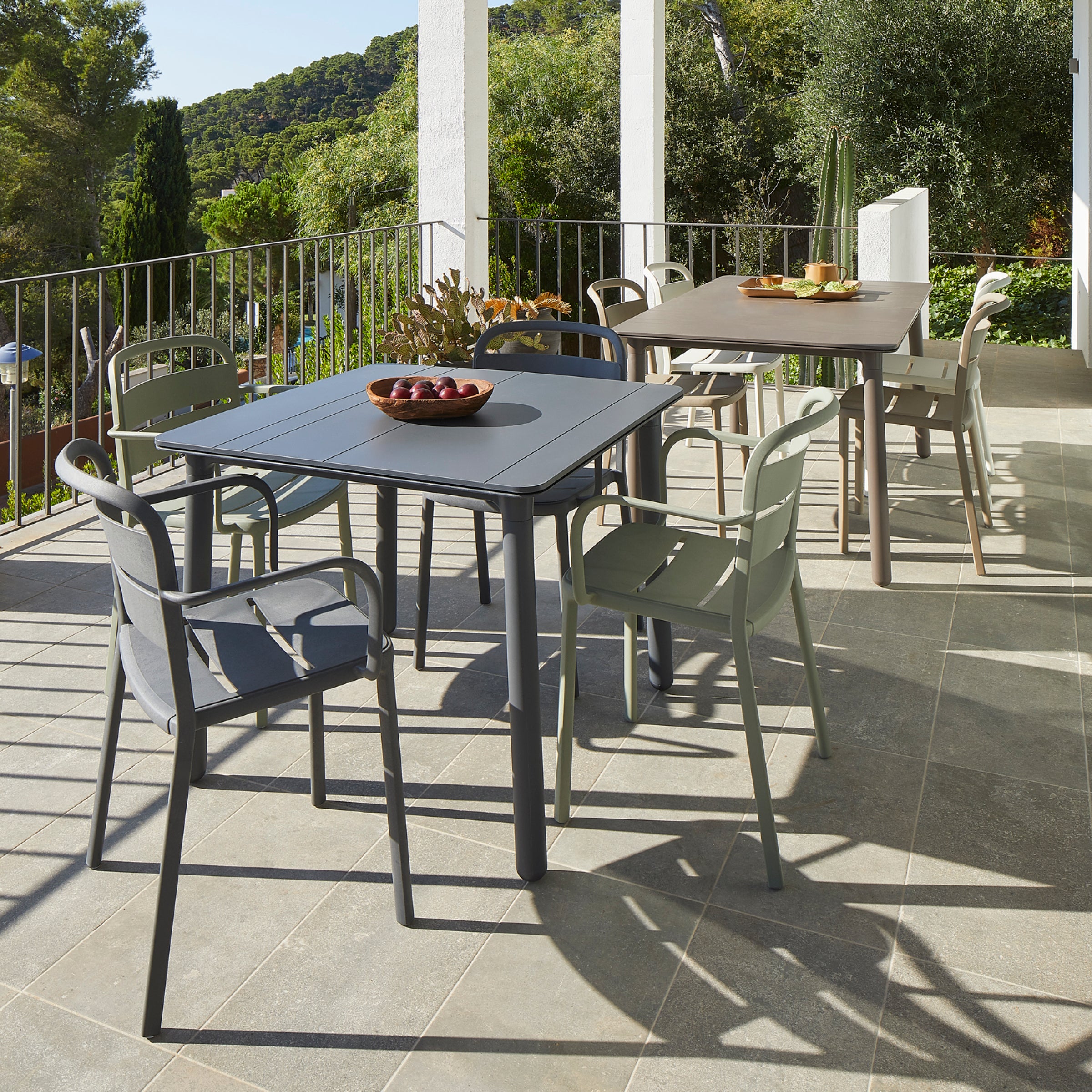 The outdoor patio boasts interior and exterior furnishings, including two tables with Resols Como Chairs under a covered terrace. A bowl of fruit decorates one table. Encircled by trees, hills in view, sunlight casts shadows on the tile floor, merging nature with sustainable design.