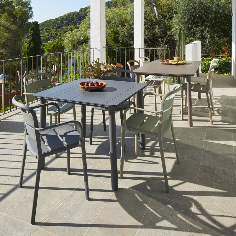 The outdoor patio boasts interior and exterior furnishings, including two tables with Resols Como Chairs under a covered terrace. A bowl of fruit decorates one table. Encircled by trees, hills in view, sunlight casts shadows on the tile floor, merging nature with sustainable design.