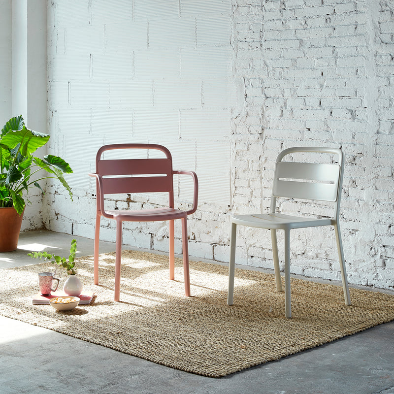 Two Resol Como Chairs, one pink and one white, stack on a woven rug in a bright room with white brick walls. A green plant in a brown pot sits nearby, along with a small coffee setup against the wall.