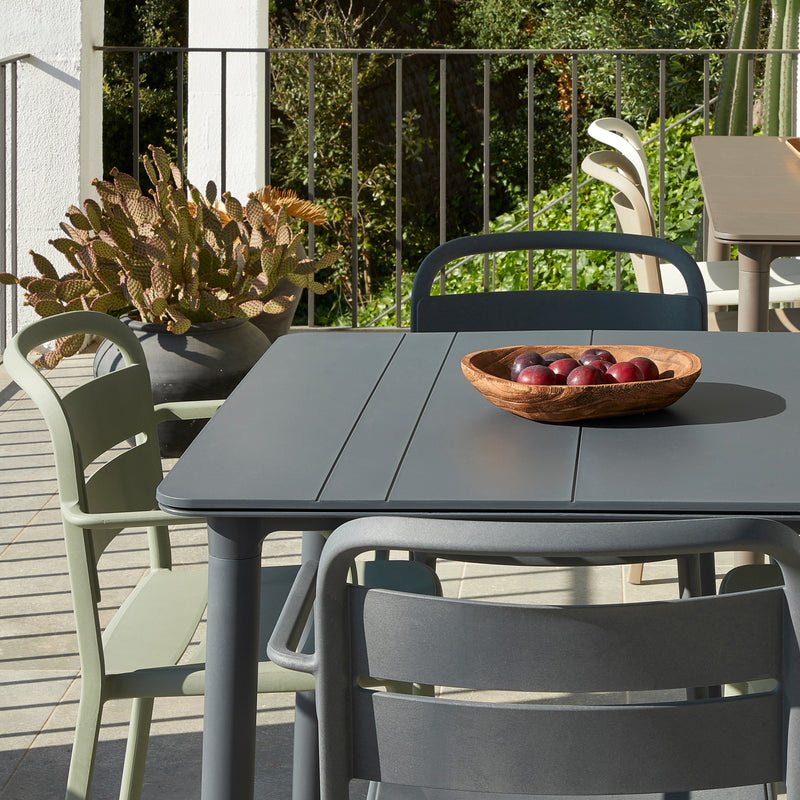 The outdoor patio scene features interior and exterior furniture including a gray table and a wooden bowl of red apples. Resols Como chairs, stackable in shades of green and gray, encircle the table, while a potted cactus and lush greenery enhance the natural, sustainable setting.