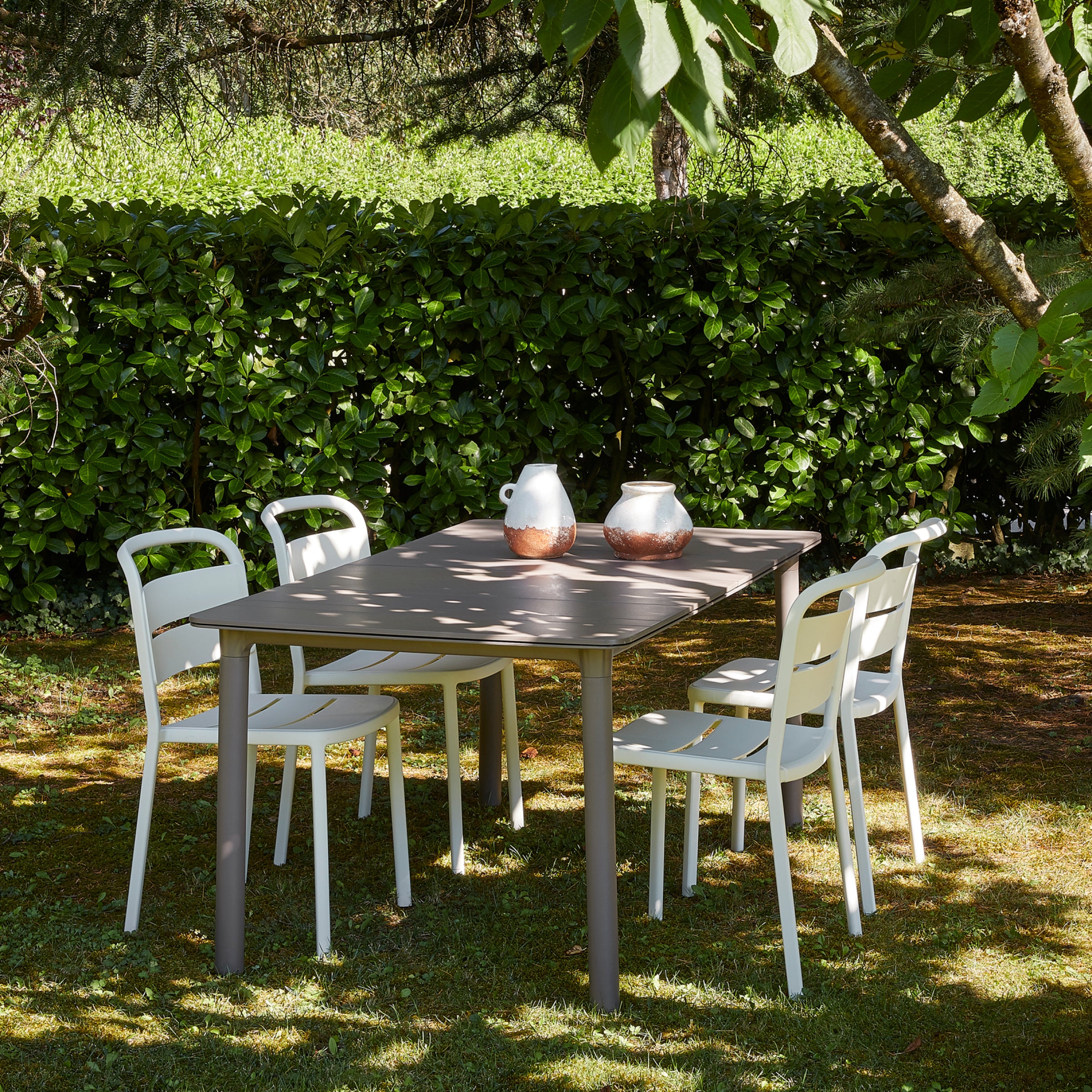 A sustainable outdoor patio set features a rectangular table and four white stackable Resol Como Chairs on grass under trees. Two white and copper pitchers sit on the table, backed by a leafy green hedge.