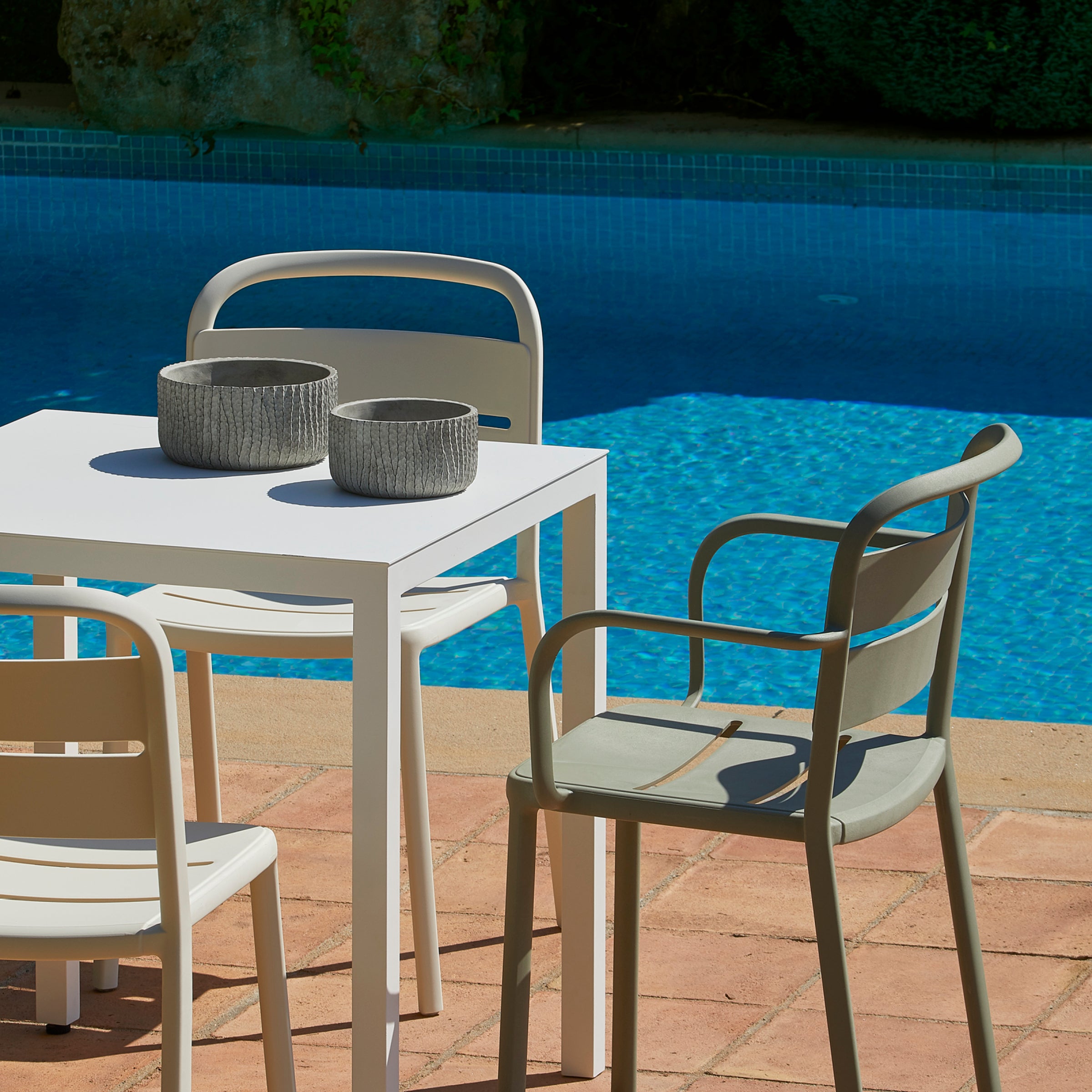 An outdoor patio scene features a white square table with four Resol Como Chairs on terracotta tiles. Two textured gray bowls rest on the table. The clear blue swimming pool is bordered by green shrubs, as bright sunlight casts shadows over this sustainable production ensemble.