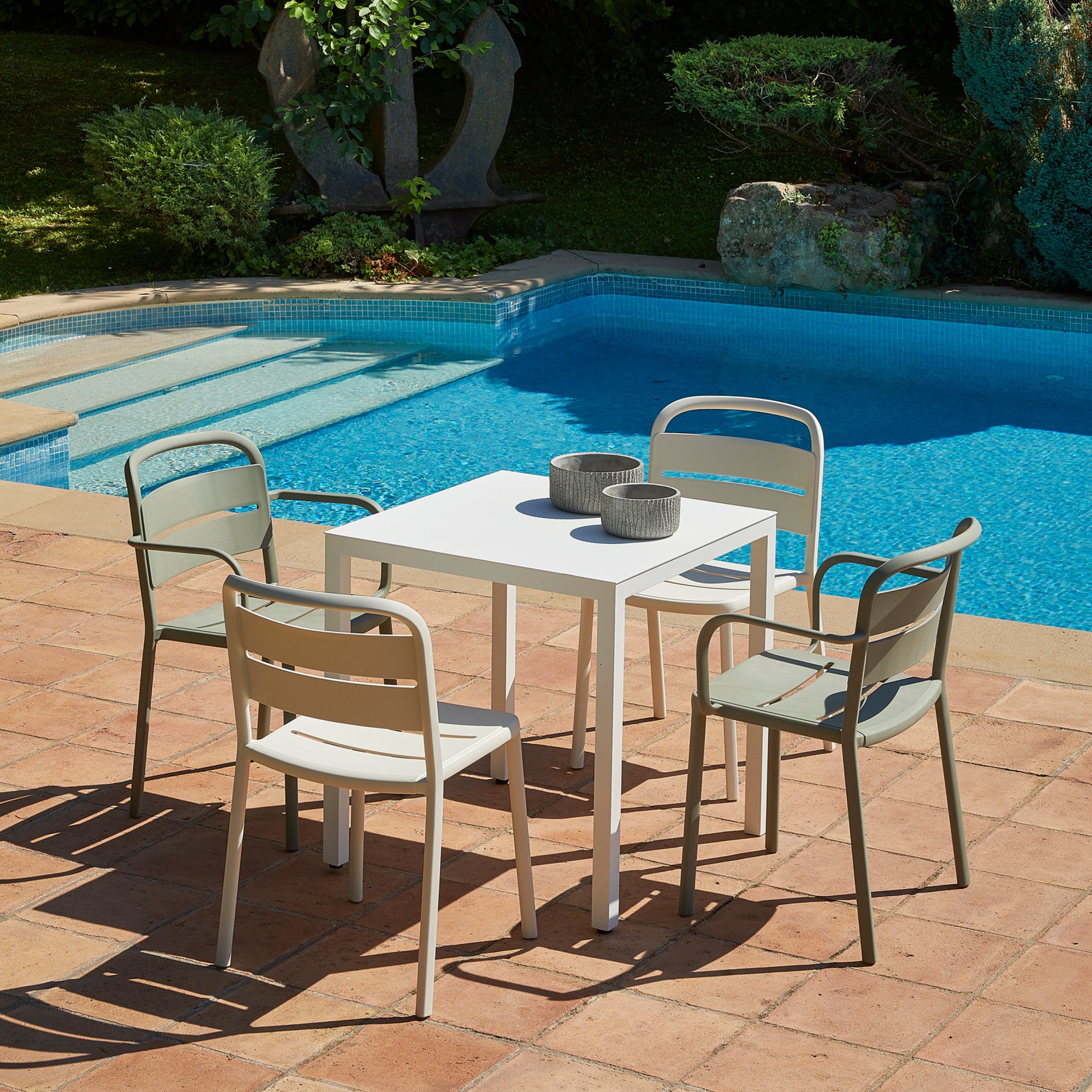 A square white table is surrounded by four Resol Como Chairs on a terracotta tile floor, with two small bowls placed on the table. Crafted sustainably, the set sits beside a clear blue pool with lush green plants in the background.