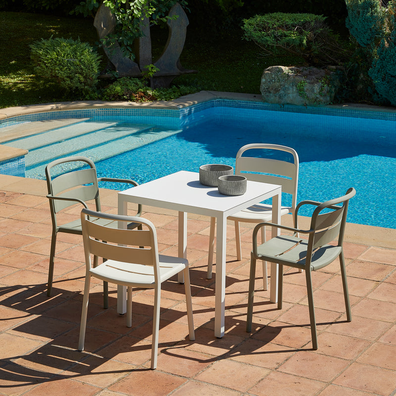 A square white table is surrounded by four Resol Como Chairs on a terracotta tile floor, with two small bowls placed on the table. Crafted sustainably, the set sits beside a clear blue pool with lush green plants in the background.