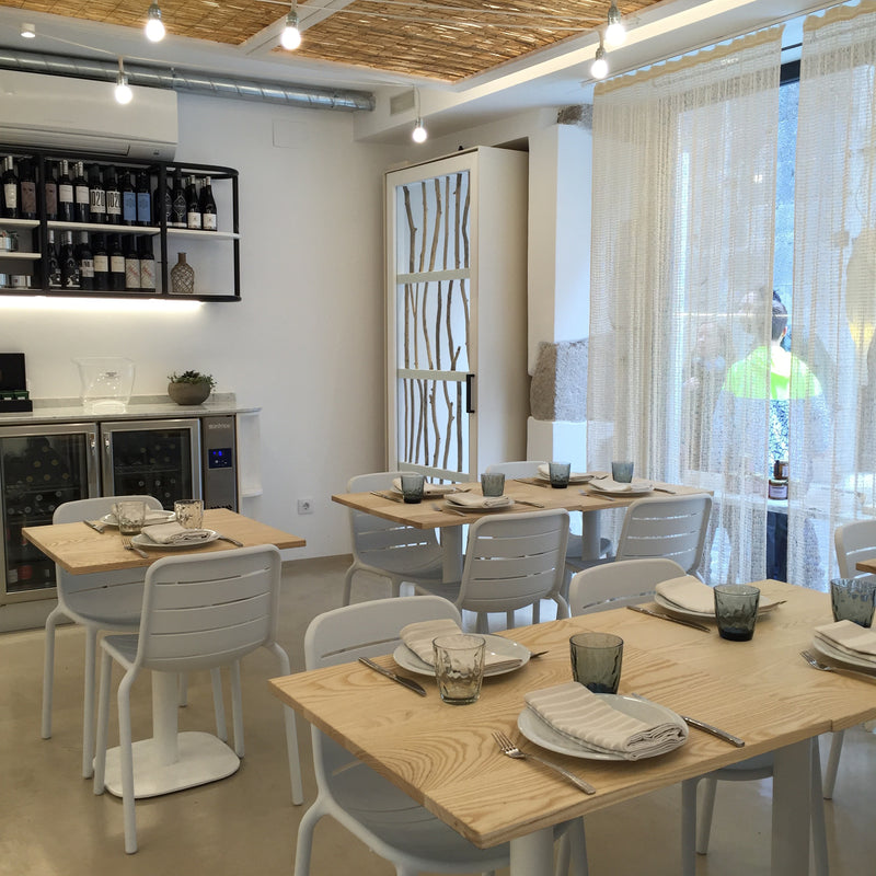 The dining area boasts UV-protected wooden tables with plates, glasses, and napkins. Surrounding the tables are Resols stackable Gina Chairs in white. On the left is a wine shelf and a small fridge, while sheer curtains let sunlight flood the room through a large window.