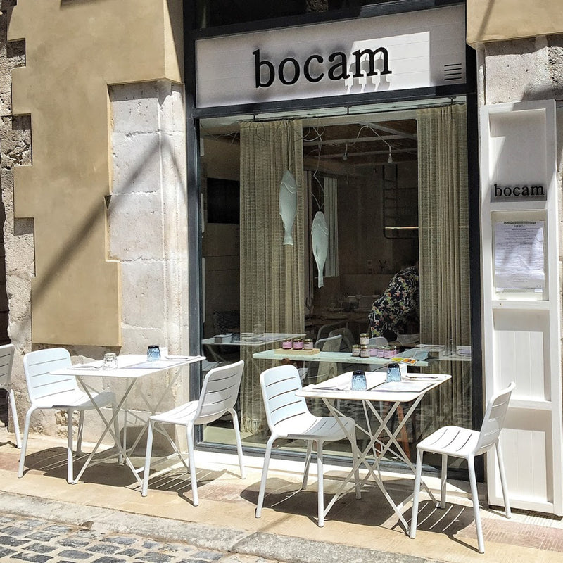 The outdoor seating of the cafe bocam features UV-protected, white tables and Resols stackable Gina Chairs. The glass-fronted entrance reveals an interior with hanging lights and curtains. A menu stands outside, as sunlight casts shadows on the cobblestone pavement.