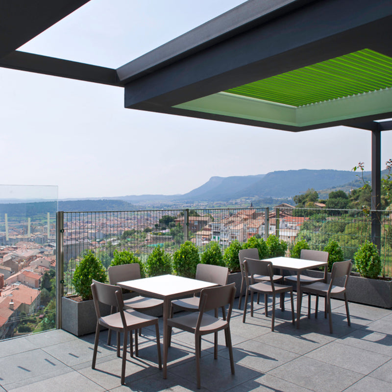 A rooftop terrace with Resols Lisboa Armchairs offers views of a scenic town and hills. Shaded by a pergola, potted shrubs line the glass railing. The UV-protected furniture, including modern tables and chairs, stays pristine under the open sky.